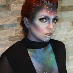 Female model with red hair in up-do of french braid down middle and glitter accents with alien makeup on face