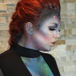 Female model with red hair in up-do of french braid down middle and glitter accents with alien makeup on face